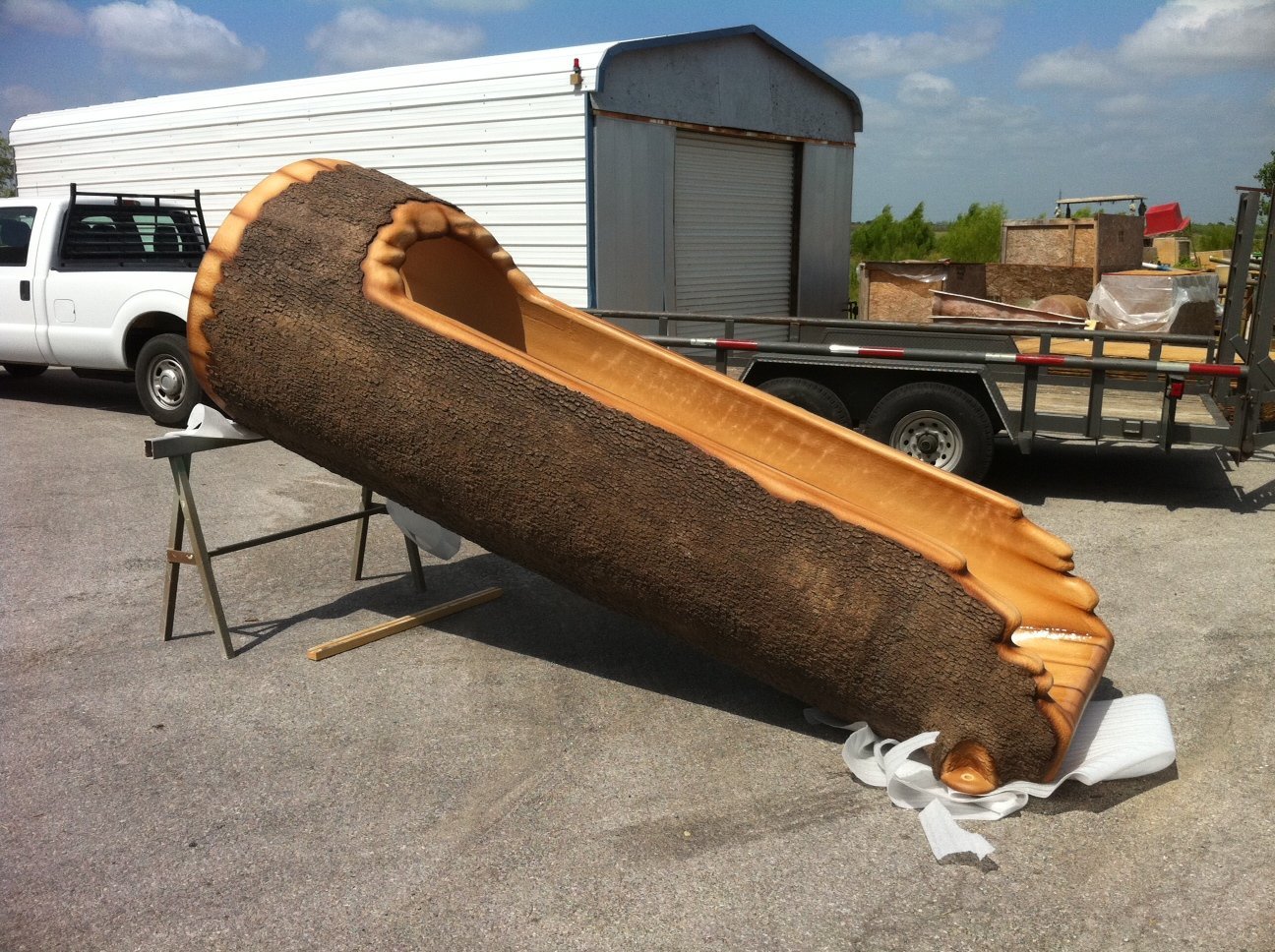 Fabricated log element lying on sawhorses, showing bark texture outside and a molded slide channel inside.